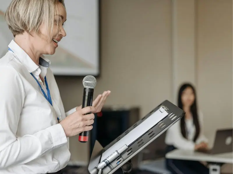 A professional woman confidently speaks at a podium, holding a microphone while presenting to an audience.