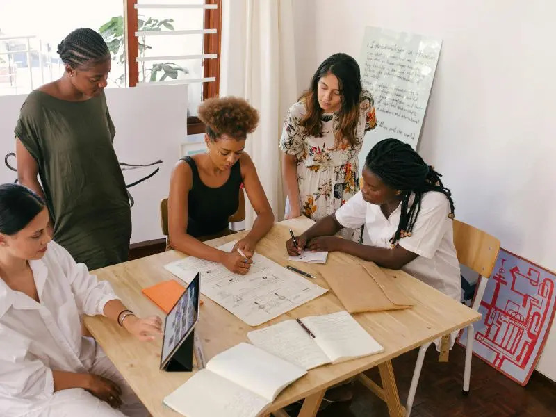 A group of female professionals working together in an office, contributing to a project by discussing ideas, writing on a dry erase board, and using a laptop.